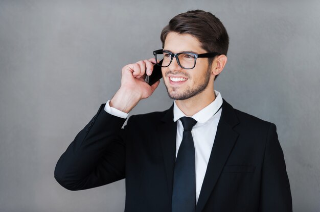 Good business talk. Happy young businessman talking on the mobile phone and looking away while standing against grey background