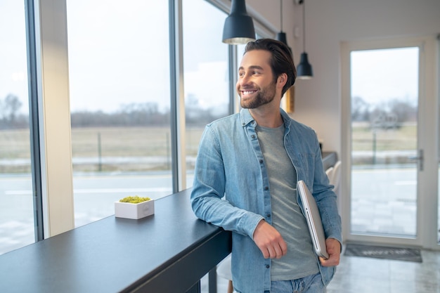 Good day. Smiling confident man standing with laptop looking out window indoors during day