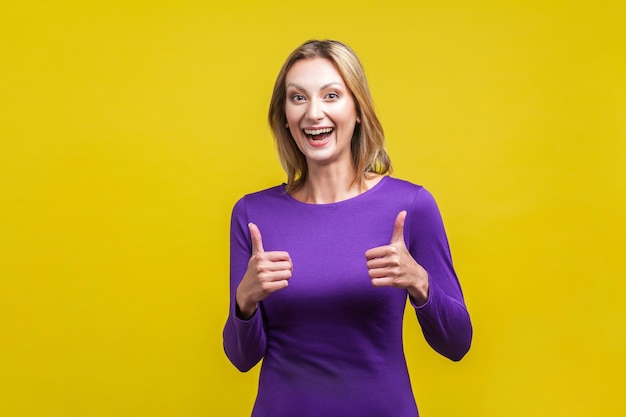 Good job Portrait of extremely happy woman in tight purple dress showing thumbs up and smiling broadly at camera overjoyed excited expression indoor studio shot isolated on yellow background