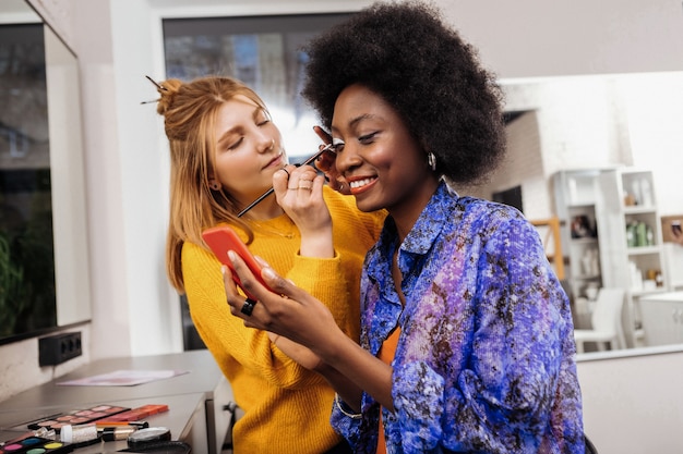 Good job. Pretty young stylist in a yellow thick sweater looking concentrated while putting silver eyeshadows on models eyelids