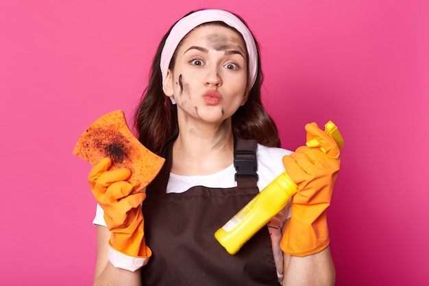 Good looking adorable woman with dirt on her face protruding her lips, holding bottle of cleaning liquid