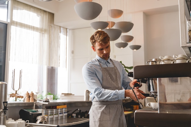 Good-looking barman in apron pouring hot beverage from the tap of coffee machine into cup