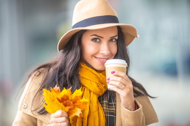 Good-looking female holds a cup of takeaway coffee and a bunch of picked colorful leaves.