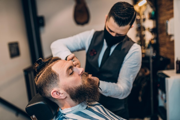 Good looking hipster young bearded man visiting hairstylist in barber shop. Barber is wearing face protective mask due to Coronavirus.