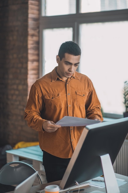 Good looking intelligent man holding the document while standing near his desk