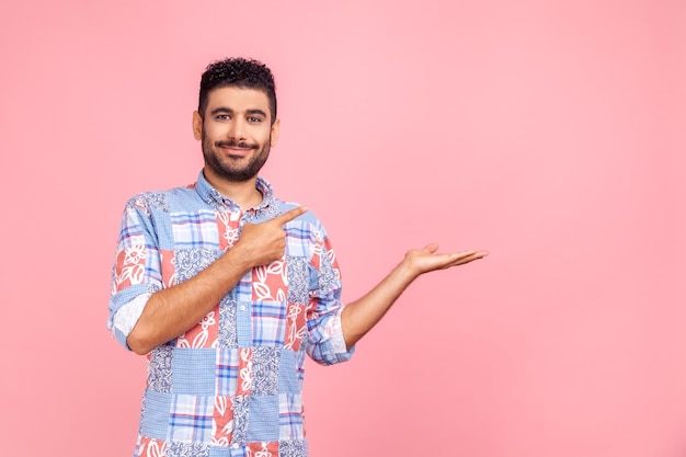 Good looking joyful man with beard in blue shirt presenting advertising area on her palm and pointing to copy space holding empty place for commercial Indoor studio shot isolated on pink background