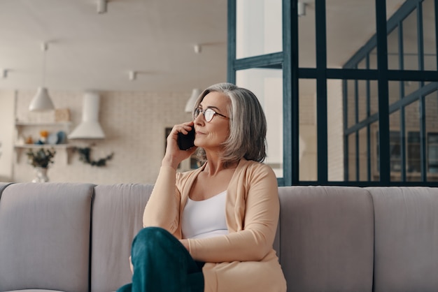 Good looking senior woman in casual clothing talking on the smart phone while sitting on the sofa at home