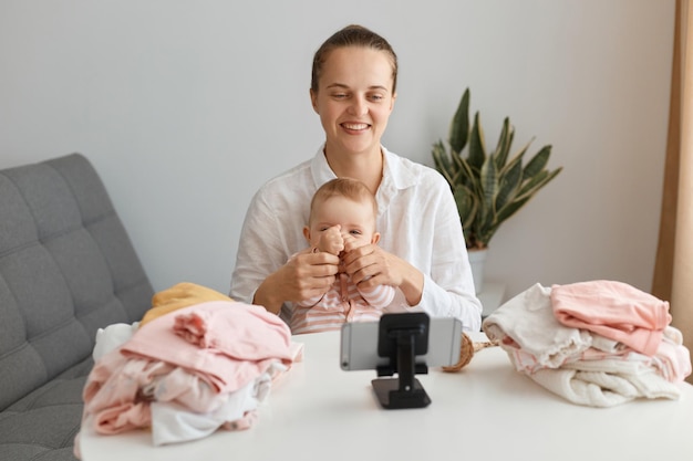 Good looking young adult woman blogger and her daughter sitting in front of phone on tripod and shooting video with sale of children's things, smiling happily while talking with followers.