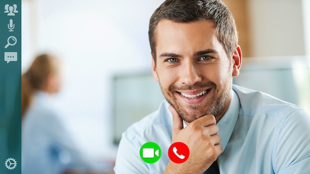 Good looking young man in formalwear looking at camera and keeping hand on chin while having video call