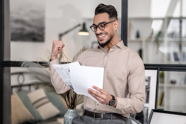 Good moments. Rejoicing young adult elegant man holding documents standing in room at home