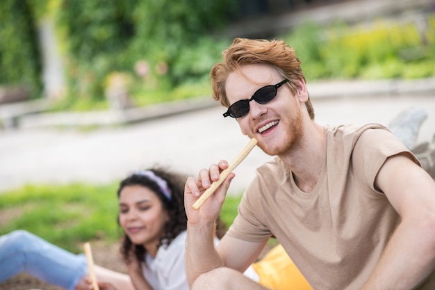 Good mood. Red-haired happy guy in sunglasses and light tshirt eating grissini on picnic on fine day