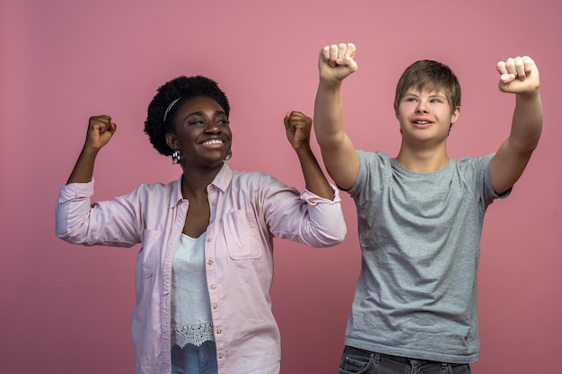 Good mood. Smiling dark-skinned woman looking at caucasian guy with down syndrome standing with raised fists on light background