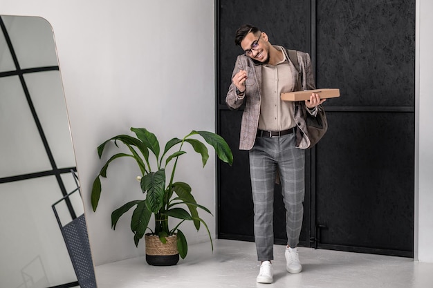 Good mood. Smiling young bearded man in suit with glasses holding box talking on smartphone at home