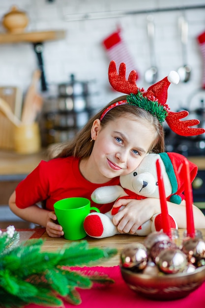 Good morning. A cute sleepy girl drinks tea at the kitchen table and hugs a teddy bear sitting in the kitchen.