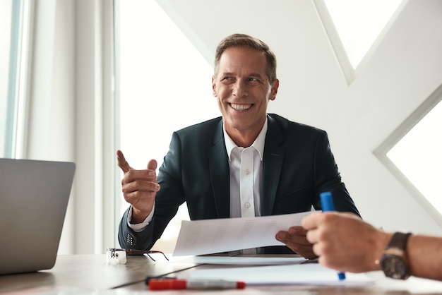 Good news. Handsome and cheerful mature man in formal wear gesturing and smiling while sitting in the office. Business people. Business meeting