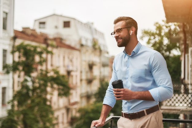 Good news young and handsome bearded man in eyeglasses and headphones holding cup of coffee