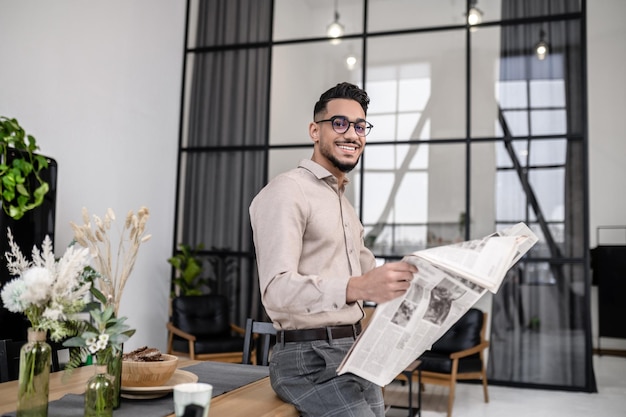 Good start. Young bearded attractive man in glasses with newspaper smiling at camera standing leaning on table in cozy bright room