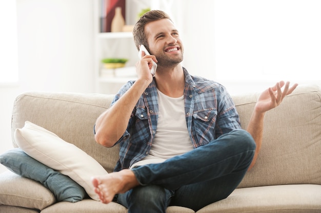 Good talk with friend. Cheerful young man talking on the mobile phone and gesturing while sitting on sofa