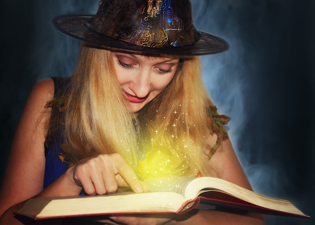 Good witch in the hat reads magic spells in the book on the fog background.