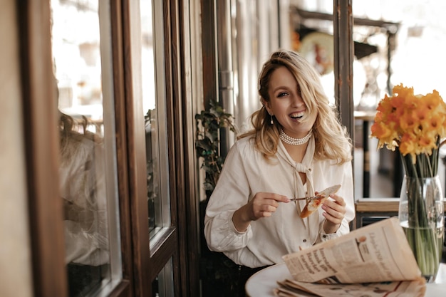 Goodhumored young blonde woman in white blouse and stylish pearl necklace laughs holds knife and piece of bread and sits in restaurant