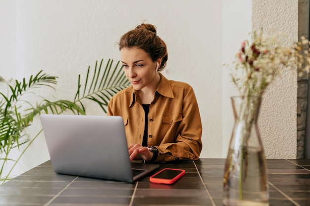 Goodlooking stylish young lady with collected hair and nude make up wearing shirt typing on laptop in cafe Concept of use and working remote