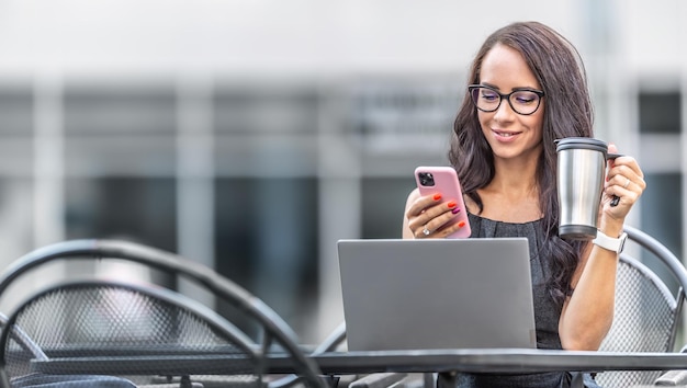 Goodlooking woman drinks coffee from reusable mug and checks text messages on a phone with an open laptop in front of her