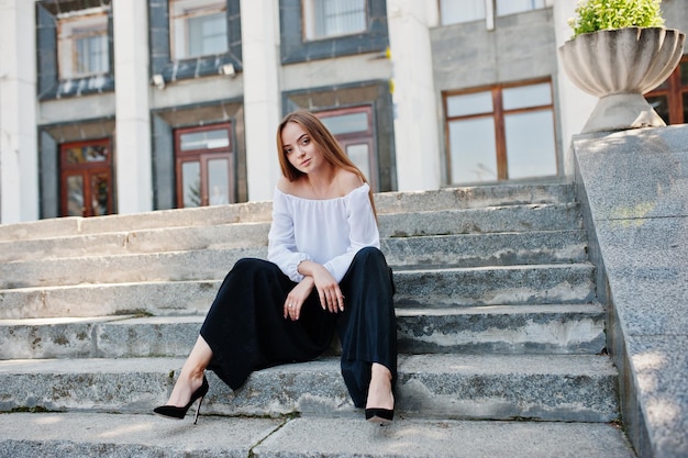 Goodlooking young woman in white blouse wide black pants and black classic high heels sitting on stairs and posing