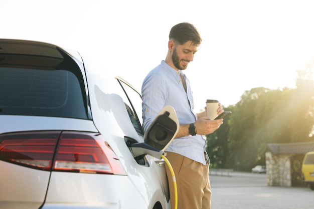 Gorgeous bearded caucasian man in a blue shirt standing near an electric car that is charging