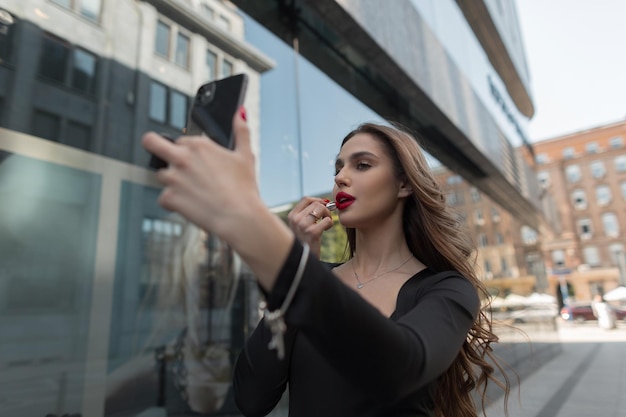 Gorgeous beautiful young fashion chic girl model paints her lips with red lipstick and looks into a smartphone in the city