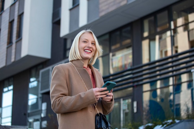 Gorgeous beautiful young woman with blonde hair messaging on the smart-phone at the city street background. pretty girl having smart phone conversation