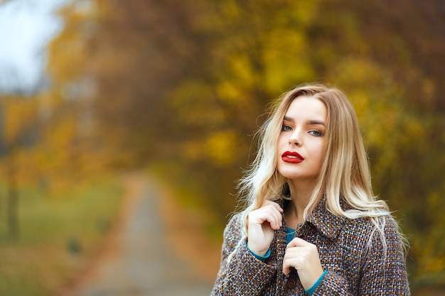 Gorgeous blonde girl with red lips wearing grey coat posing in autumn park. Empty space