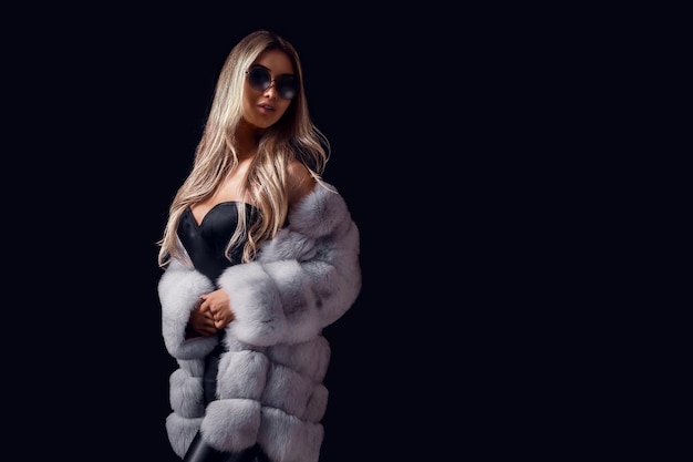 Gorgeous blonde woman posing in luxurious fur coat and sunglasses while standing on black background