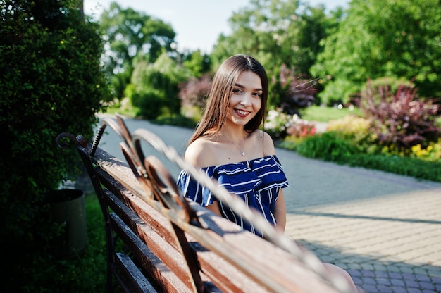 Gorgeous brunette woman sitting on bench at street of city wear on blue striped dress.