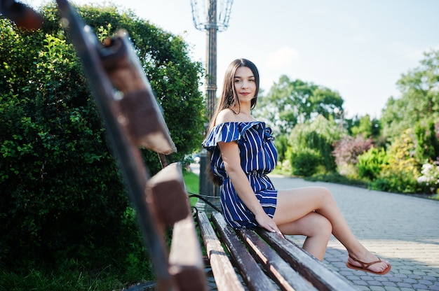 Gorgeous brunette woman sitting on bench at street of city wear on blue striped dress.