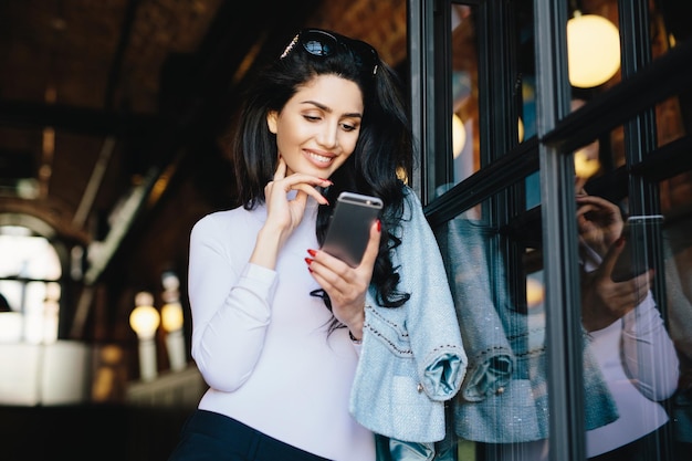 Gorgeous brunette woman in sunglasses dressed formally using cell phone flirting with handsome man on internet waiting for his answer looking at screen with pleasure and great joy sitting in cafe