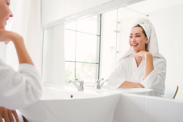 Gorgeous confident middleaged adult woman wearing a towel on her head sitting and smiling in front of the mirror in the home bathroom A pretty attractive woman beauty lifestyle and cosmetic concept