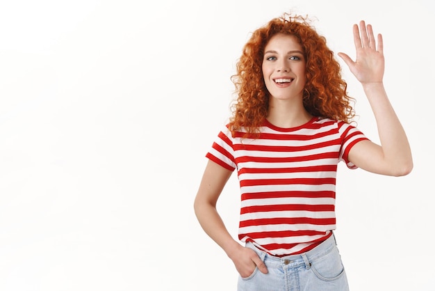 Gorgeous curlyhaired caucasian redhead woman raise palm high five smiling amused lively greeting waving hand hello hi whats up gesture standing relaxed selfassured white background