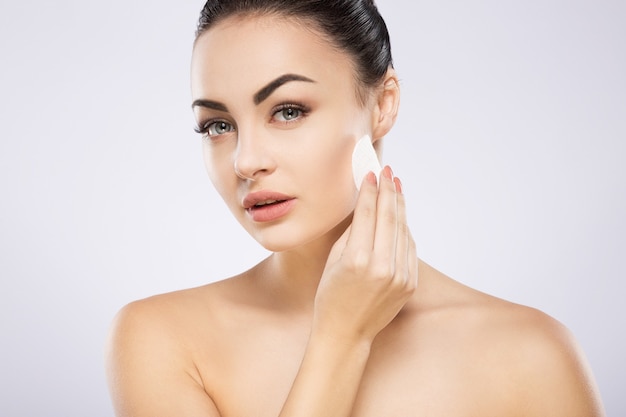 Gorgeous girl with black hair fixed behind, big eyes, thick eyebrows and naked shoulders standing at gray  wall and holding cosmetic cleaning sponge, copy space.