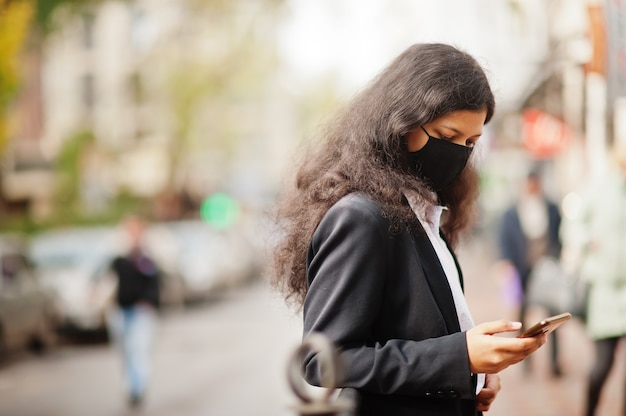 Gorgeous indian woman wear formal and black face mask, posing at street during covid pandemia, watching news on her smartphone.