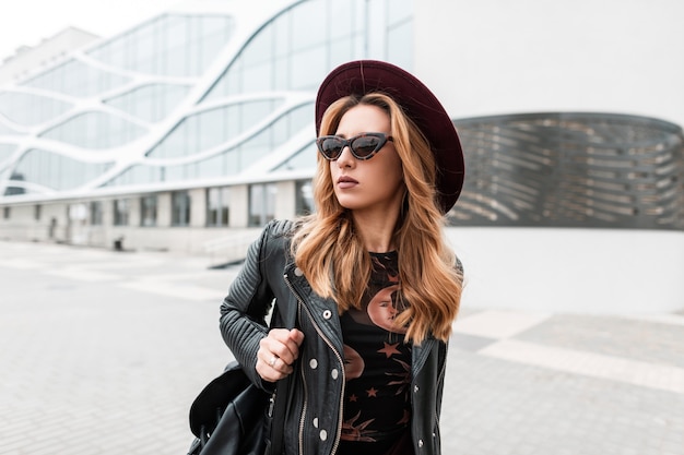 Gorgeous red-haired hipster young woman in dark sunglasses in an elegant hat in a stylish black leather jacket