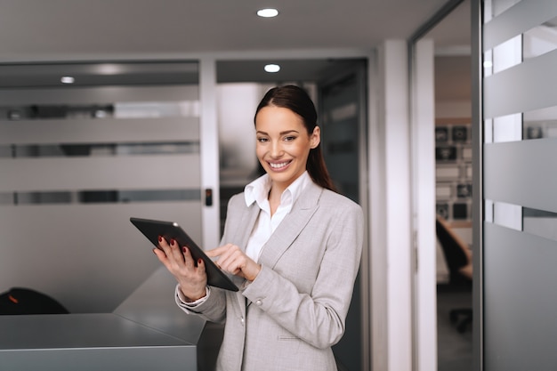 Gorgeous smiling brunette in formal wear using tablet. Corporate firm interior. Don't be busy, be productive.