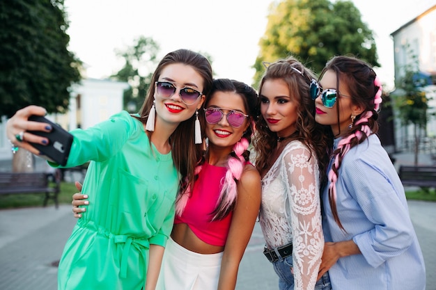 Gorgeous smiling women in sunglasses posing at street and tacking self portrait at smartphones and call phones. Fashionable girls with pink brides wearing in colorful clothes tacking photo. Fashion.