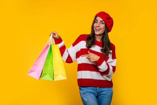 Gorgeous stylish woman in a striped sweater and red beret with many colorful shopping bags in hands is posing isolated on yellow background