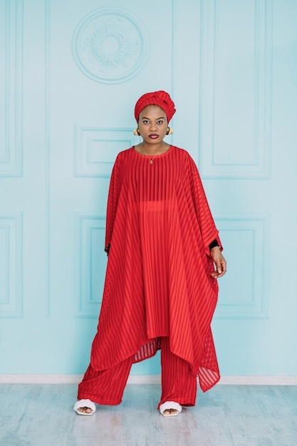 Gorgeous trendy young African American woman in red stylish suit and headscarf, standing against light blue studio wall background.