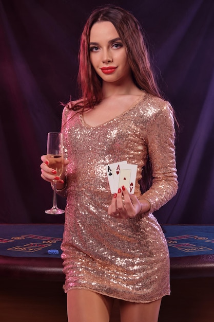 Gorgeous woman in golden dress is showing two cards holding champagne while posing at playing table in casino Black background colorful backlights Gambling entertainment poker Closeup