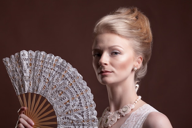 Gorgeous woman holding an oriental fan in hands on brown background in studio photo. Rococo style. Vintage and glamour