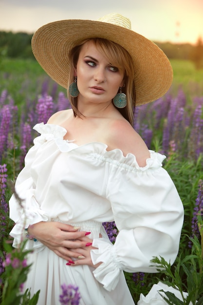 A gorgeous woman in a long light summer dress and hat on a field of flowers