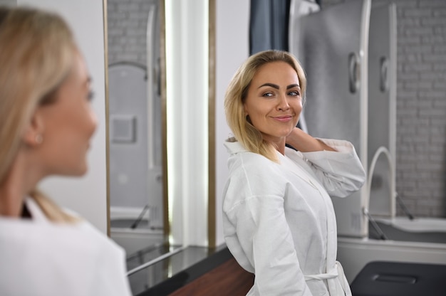 A gorgeous woman smiles looking at her mirror image. Woman in spa resort center