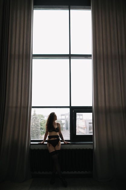 Gorgeous woman wearing black lingerie indoors Portrait of a beautiful dreamy girl in a black underwear by the window