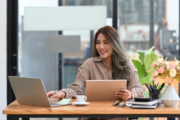 Gorgeous young businesswoman holding digital tablet and using computer laptop